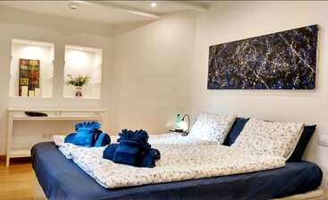 Un dormitorio acogedor y moderno con una cama de matrimonio, ropa de cama de flores, cisnes de toalla azul, arte abstracto en la pared, un escritorio blanco y una iluminación suave y cálida.