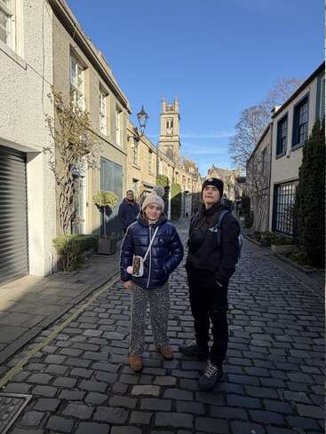 Two people in winter clothes stand on a cobblestone street, with another person in the background. Historic buildings and a church tower are visible under a clear sky.