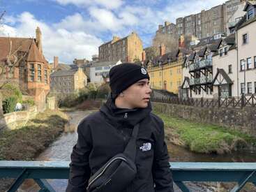A person wearing a black jacket and beanie stands on a bridge, with picturesque old European buildings and a small river in the scenic background.