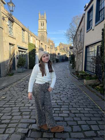 A girl poses on a quaint cobblestone street lined with charming buildings, wearing a white sweater, animal print pants, and a beanie under a clear blue sky.