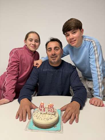 A man is celebrating his 45th birthday with a cake, accompanied by two smiling children, one girl in pink and one boy in blue.