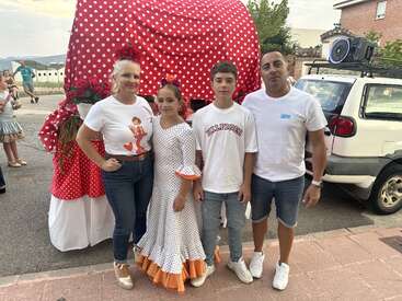 A family of four poses together outdoors. The mother and daughter wear festive attire, while the father and son dress casually. Behind them, a polka-dotted cart.