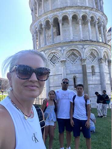 A family poses for a selfie in front of the Leaning Tower of Pisa on a sunny day, wearing casual summer clothes and sunglasses, enjoying their vacation.