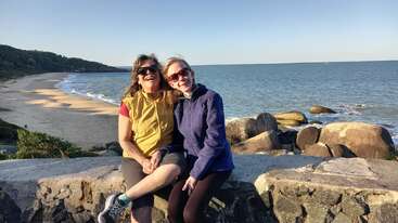 Dos mujeres sentadas en un muro de piedra junto al océano, sonríen y llevan gafas de sol. Detrás de ellas hay una playa de arena, rocas y un cielo azul despejado.