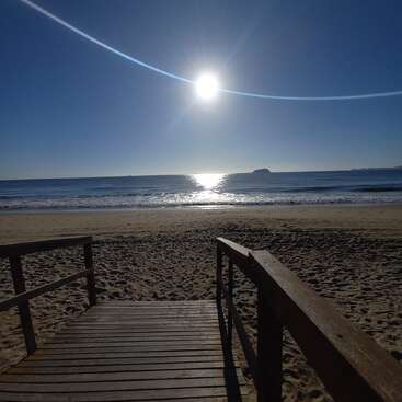 Un paseo marítimo de madera conduce a una playa de arena bajo un sol radiante. Olas tranquilas se encuentran con la orilla y una isla aparece en el horizonte. Un paisaje apacible.