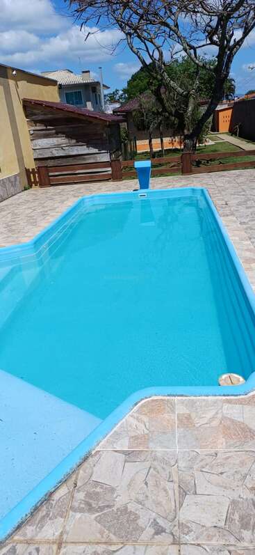 Uma piscina azul limpa com deck de azulejos, cercada por uma cerca, árvores e edifícios sob um céu ensolarado, criando uma atmosfera relaxante ao ar livre.