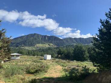 A beautiful landscape features green mountains, a bright blue sky with scattered clouds, trees, and grassy fields. Two small structures sit quietly in the foreground.