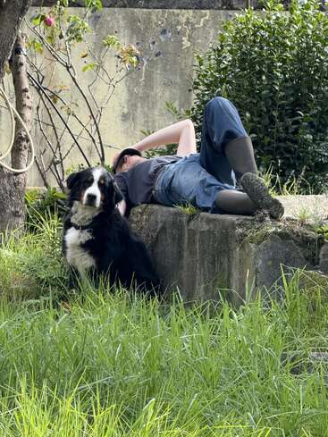 A person lies on a concrete ledge, wearing boots and jeans, relaxing with one hand over their face. A black and white dog sits nearby, alert.