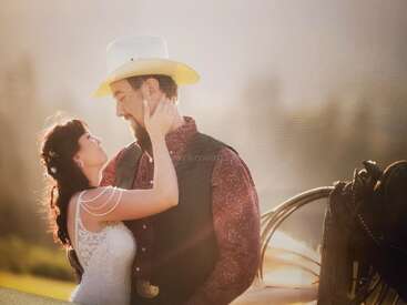 Eine Braut und ein Bräutigam umarmen sich liebevoll bei Sonnenuntergang im Freien. Der Bräutigam trägt einen Cowboyhut, die Braut ein Spitzenkleid. Warme, romantische Hochzeitsatmosphäre auf dem Land.