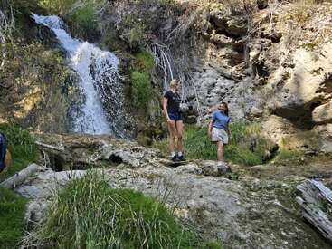 Two people stand near a beautiful rocky waterfall surrounded by green grass and foliage, smiling and enjoying a peaceful nature scene on a sunny day.