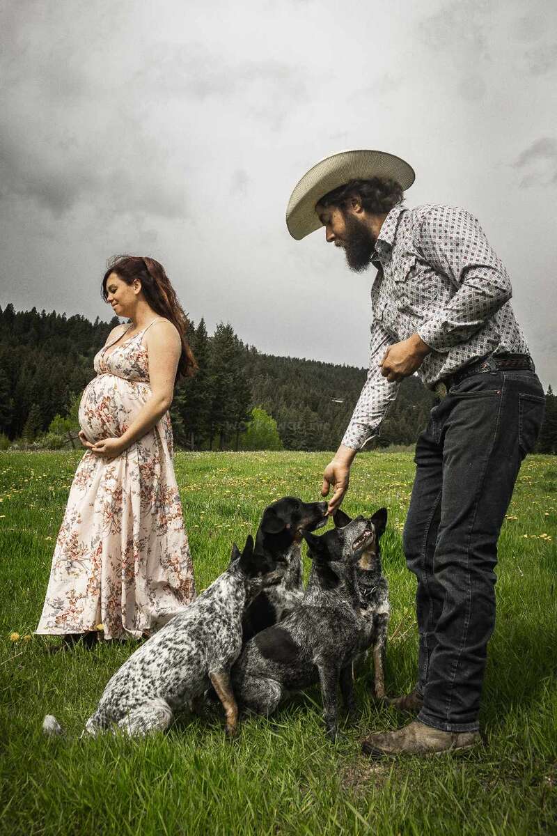 Una mujer embarazada con un vestido de flores está de pie en un campo cubierto de hierba junto a un hombre con sombrero de vaquero que da golosinas a cuatro perros atentos.