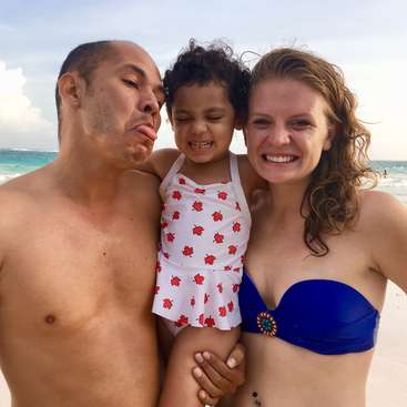 La imagen muestra a una familia de tres en una playa, con el hombre a la izquierda, la niña en el centro y la mujer a la derecha, todos sonriendo y posando para la cámara.