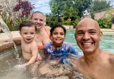 Eine fröhliche vierköpfige Familie genießt die gemeinsame Zeit in einem Swimmingpool und lächelt dabei strahlend. Üppiges Grün umgibt sie, und alle sehen entspannt und glücklich aus.
