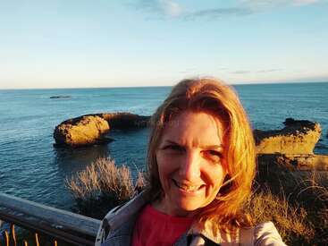 The image depicts a woman with shoulder-length blonde hair standing in front of a serene ocean view, with a large rock formation visible in the water behind her.