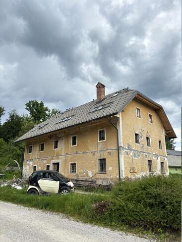 An old, weathered house with peeling yellow paint stands under a cloudy sky. A small black and white car is parked outside on overgrown grass.