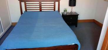 Uma cama bem arrumada com uma capa azul fica em um quarto simples. Há uma cabeceira de madeira, uma mesa de cabeceira preta, uma lâmpada, uma pequena planta e uma decoração neutra.