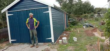 Um homem está em frente a um galpão azul recém-pintado, cercado por grama, latas de tinta, canteiros de jardim, ferramentas e vários itens espalhados pelo quintal.