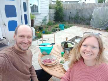 Um homem e uma mulher sorriem em uma mesa ao ar livre, desfrutando de uma refeição. O cenário é um quintal com uma mesa de madeira, um jardim e itens de comida visíveis.