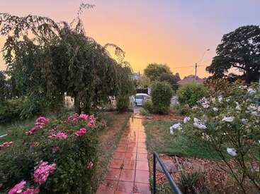 Un chemin de jardin serein bordé de fleurs roses et blanches, d'une végétation luxuriante et d'arbres, mène à une allée au coucher du soleil, avec un ciel aux couleurs pastel au-dessus.