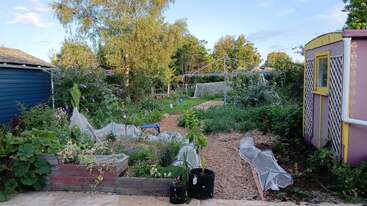 A lush backyard garden with raised beds, protective netting over plants, trees, a pathway, a blue bench, garden tools, and a rotary clothesline under clear skies.