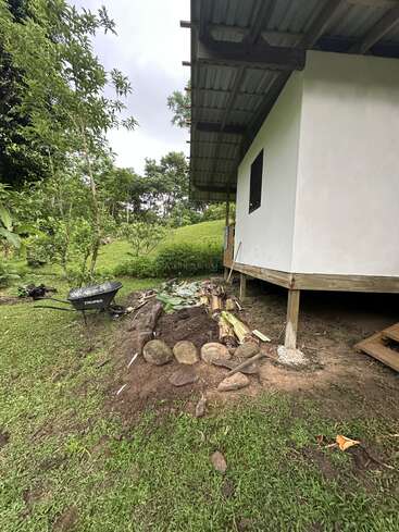 Ein kleines weißes Haus auf Stelzen steht neben einem Haufen Erde, Steinen und Bananenblättern in einem üppigen, grünen, ländlichen Gartengebiet mit Bäumen.