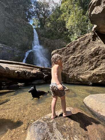 Ein kleines Kind steht barfuß auf einem Felsen an einem klaren Teich, mit einem schwarzen Hund in der Nähe. Hinter ihnen stürzt ein hoher Wasserfall in üppiges Grün.