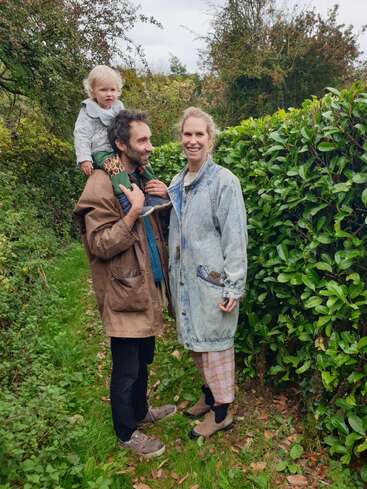 Una familia feliz se encuentra al aire libre en un camino verde y frondoso. Un hombre lleva a un niño pequeño a hombros mientras una mujer sonríe junto a ellos, disfrutando de la naturaleza.