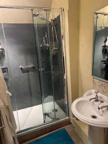 Esta imagen muestra un cuarto de baño compacto con una ducha acristalada, un lavabo de pedestal, un gran espejo y una alfombrilla azul, con paredes de color beige.