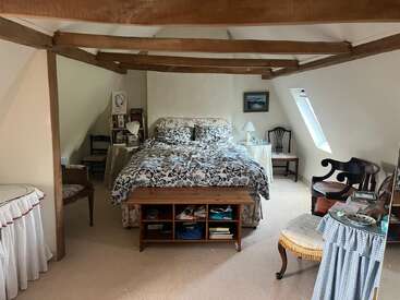 Esta acogedora habitación abuhardillada cuenta con vigas de madera a la vista, una cama doble con ropa de cama de flores, mesillas de noche, sillas de época, libros y una claraboya, creando un refugio encantador.