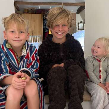 Tres niños sonrientes están sentados juntos dentro de casa. El niño de la izquierda lleva una bata a rayas, el del medio lleva un traje marrón peludo y el más pequeño ríe alegremente. Ambiente acogedor.