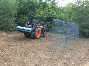 The image depicts a blue and orange tractor situated in a dirt clearing, with a metal fence in the background and trees surrounding the area.
