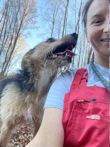 The image shows a woman in red overalls taking a selfie with a dog in a wooded area, surrounded by trees and dry leaves on the ground.