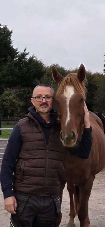 Ein Mann mit Brille und brauner Weste steht dicht neben einem braunen Pferd und hält ihm sanft das Gesicht. Im Hintergrund sind Bäume und ein bewölkter Himmel zu sehen.