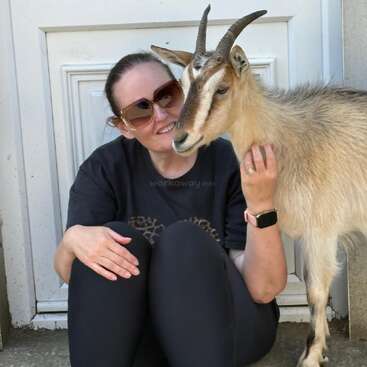 A woman wearing sunglasses and a dark shirt sits on the ground, smiling and cuddling a friendly goat. They appear happy, relaxed, and affectionate together.