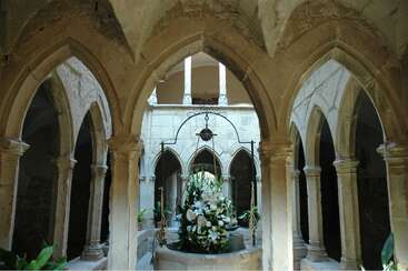 Cette image montre un cloître médiéval en pierre, avec des arcs en ogive, des colonnes et un puits central orné de fleurs blanches et de verdure, créant une atmosphère sereine et historique.