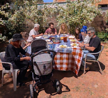 Une famille est réunie en plein air autour d'une table garnie de plats, partageant un repas sous les fleurs épanouies. On y voit six adultes, deux enfants et une poussette. L'ambiance est chaleureuse et ensoleillée.