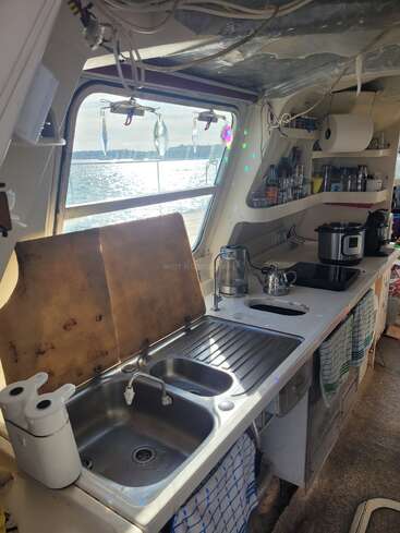 Eine kompakte Bootsküche mit Doppelspüle, Einbaugeräten, hängenden Küchenutensilien und Regalen. Durch ein Fenster mit Blick aufs Wasser strömt Sonnenlicht herein und sorgt für eine helle Atmosphäre.