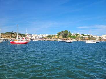 Strahlend blaues Wasser, auf dem mehrere Segelboote friedlich dahintreiben. Ein rotes Boot sticht besonders hervor. Sonniger Himmel, grüne Hügel und weiße Gebäude an der Küste runden dieses idyllische Bild eines Küstendorfes ab.