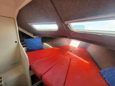 Dieses Bild zeigt das gemütliche Interieur einer Bootskabine mit rot gepolsterten Liegeflächen, blauen Kissen, mit Teppich ausgekleideten Wänden, durch zwei Fenster hereinströmendem Sonnenlicht und kompakten Stauräumen.