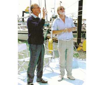 Two men stand together on a boat under sunny skies. Chairs and yachts are visible behind them. One gestures upward, both appear engaged in conversation outdoors.
