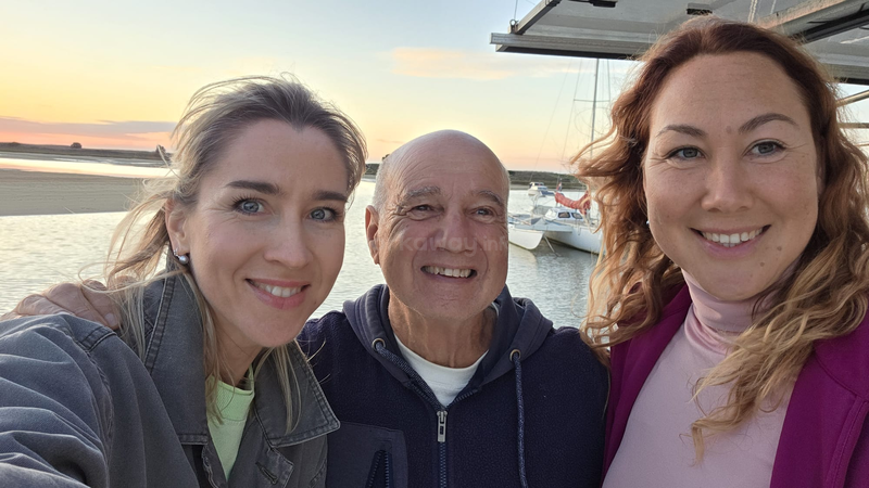 Drei Leute posieren bei Sonnenuntergang am Wasser für ein fröhliches Selfie. Hinter ihnen liegen Boote am Ufer, und der Himmel ist in warme Pastelltöne getaucht.