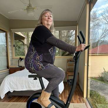 A woman is riding an exercise bike indoors, near a window with a scenic outdoor view. She is dressed comfortably and appears focused on her workout.