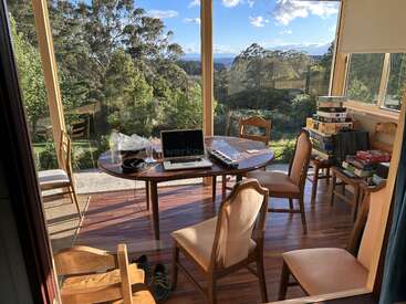 A sunlit room with large windows overlooks a lush forest. A laptop, water bottle, and keyboard sit on a wooden table surrounded by chairs and board games.