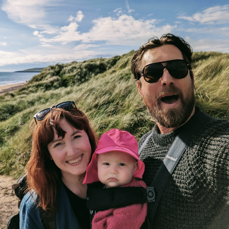 L'image représente une famille de trois personnes posant pour une photo sur une plage, avec une colline herbeuse et l'océan en arrière-plan. La famille est habillée de manière décontractée.