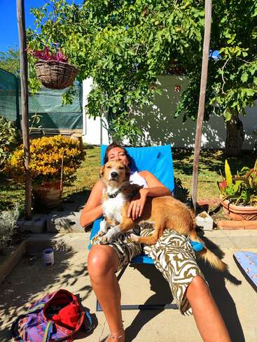 Une femme se détend sur une chaise longue bleue en plein air, souriante, avec un chien marron sur les genoux. Des arbres verts, des plantes et la lumière du soleil les entourent. Scène paisible.