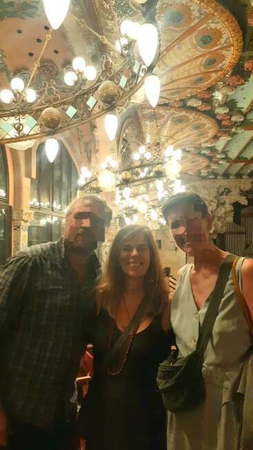 Drei Menschen mit verpixelten Gesichtern stehen in einem kunstvoll verzierten, historisch anmutenden Gebäude unter bunten Deckenleuchten und Kronleuchtern zusammen. Sie scheinen glücklich zu sein.