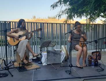 Zwei Frauen spielen im Freien Live-Musik. Die eine spielt eine Gitarre und singt, die andere spielt ein Streichinstrument. Mikrofone und Notenständer sind vorhanden.