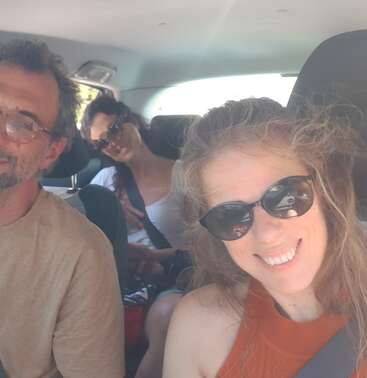 Three people are inside a car, all wearing sunglasses. The woman in front is smiling at the camera, while the others sit relaxed and comfortable. Sunlight fills the car.