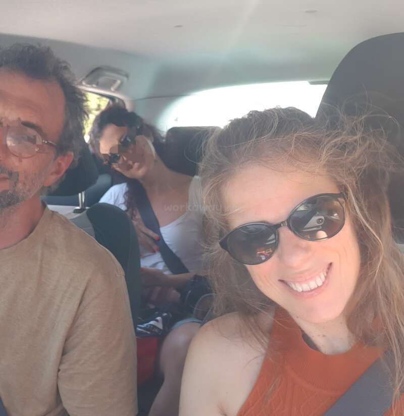 Three people are inside a car, all wearing sunglasses. The woman in front is smiling at the camera, while the others sit relaxed and comfortable. Sunlight fills the car.