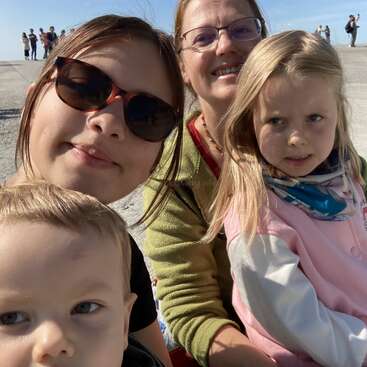 The image shows two women and two children posing for a selfie on a beach, with a crowd of people in the background under a clear blue sky.
